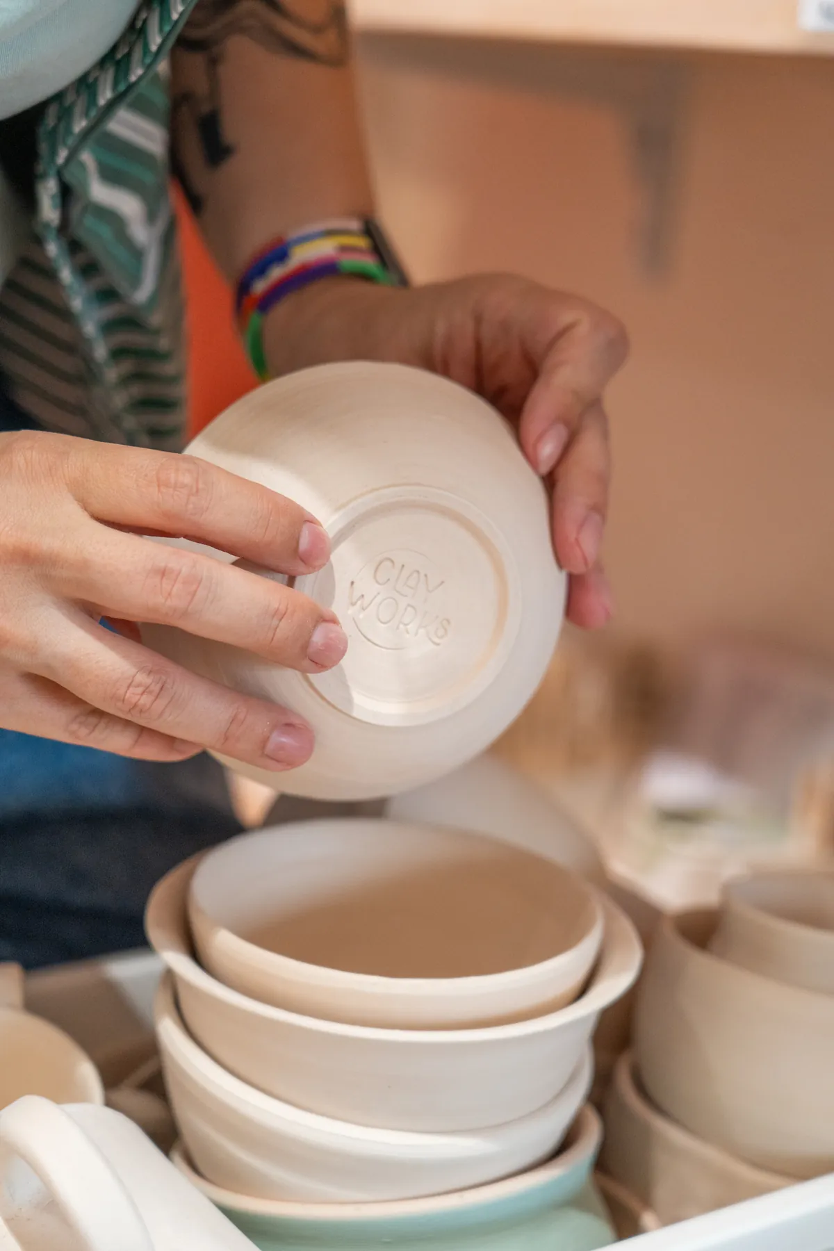 Hände halten eine helle Schale mit eingeprägtem Clayworks-Stempel über einem Stapel fertiger Schalen