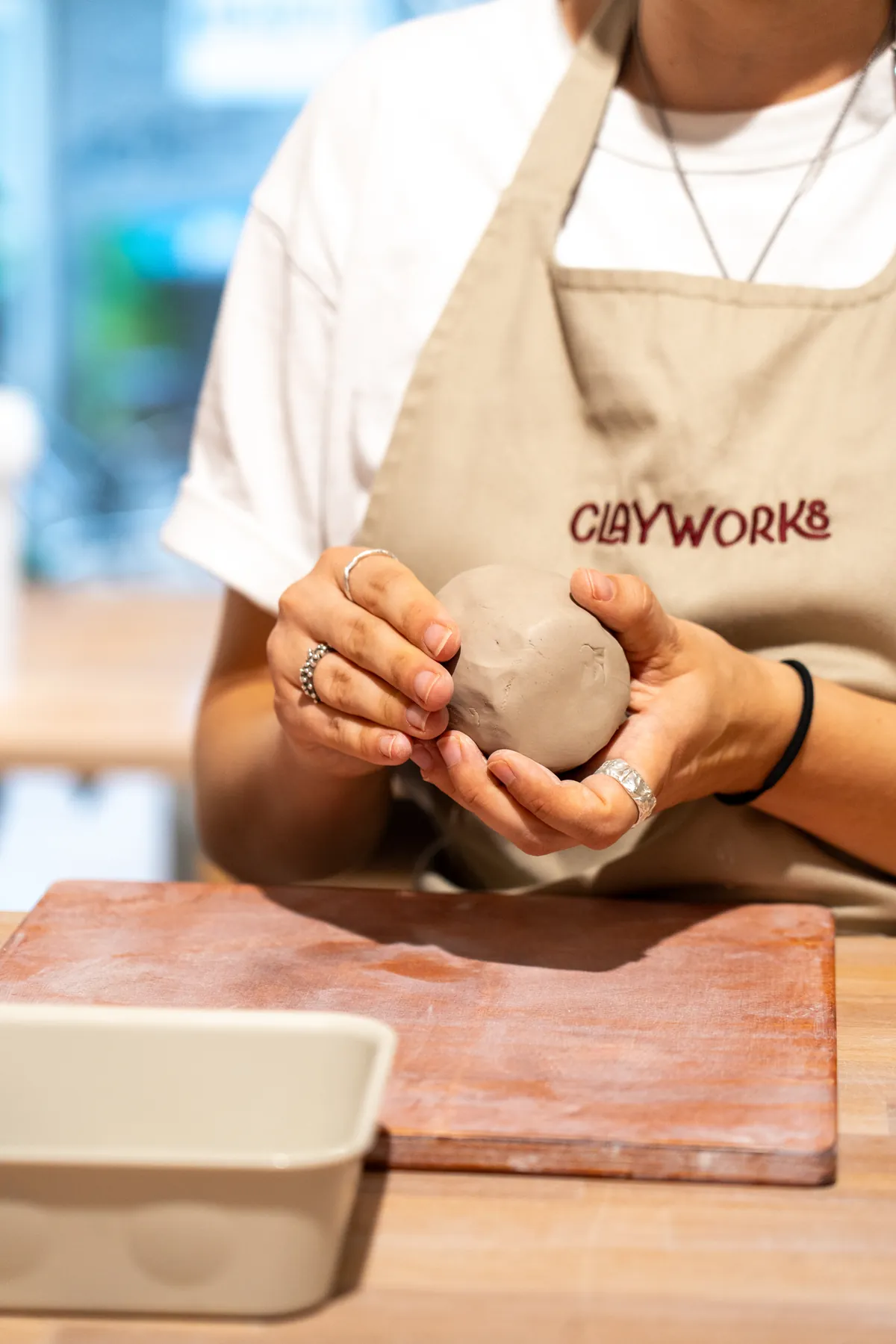 Person in Clayworks-Schürze knetet eine Tonkugel über einem Holzbrett